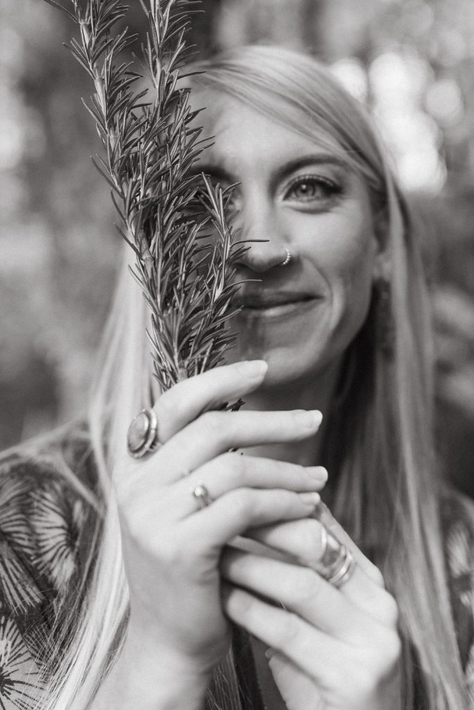 Lindsey Walker, owner of Mockingbird Remedies, in nature, holding a rosemary sprig in front of the right side of her face. Black and white photo.